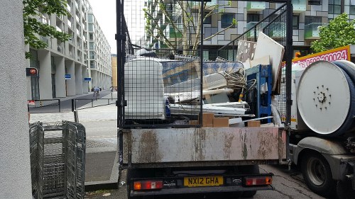 A flatbed truck parked on a city street corner in an urban area, with a black metal safety cage on the bed containing various disused items including white panels, wooden planks, and metal debris. The truck's rear end features a rusty metal bumper and is positioned near a modern residential building with white balconies. Visible in the background are a traffic light, street signs, and a pedestrian crossing, indicating an active city environment. The scene suggests a professional rubbish removal service, likely part of Garden Clearance Southgate, operating in the Southgate area, with the truck ready to collect and dispose of household or garden waste materials. The image captures the logistics of urban waste collection, emphasizing the preparation of clutter removal from nearby properties or streets, consistent with local rubbish clearance services nearby.