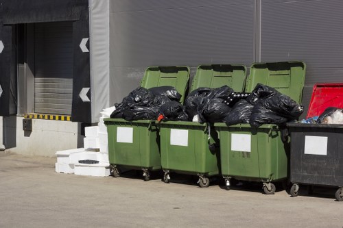 A row of four large, green wheeled rubbish skips with their lids open, filled with tightly packed black trash bags, are positioned on a paved outdoor area adjacent to a grey industrial building. To the right of the green skips is a black waste bin, also filled with rubbish, with a glimpse of red objects inside. Behind the bins, there are a few white bricks or blocks stacked on the ground near the building's wall, which features a large roller shutter door with black and yellow safety markings on its border. The environment suggests a commercial or industrial waste collection point, highlighting the type of rubbish disposal services provided by Garden Clearance Southgate for domestic or commercial clients in Southgate or nearby areas. The scene is lit by natural daylight, and the setting appears clean and organized for rubbish removal, reflecting a typical outdoor waste management setup within the local vicinity.