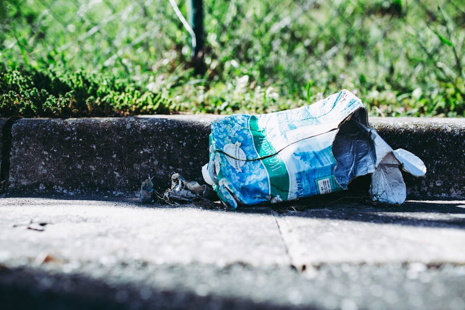 Close-up view of a large pile of crushed and crumpled aluminium cans and food packaging, predominantly in metallic silver, gold, red, blue, black, and yellow colors, scattered across a dark surface. T